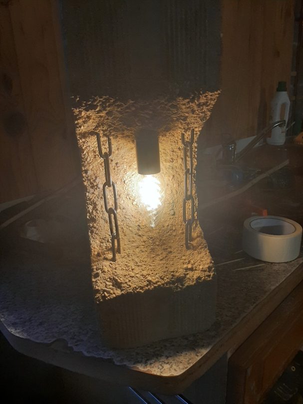 Lampe en béton avec des chaînes décoratives, éclairant une surface en bois.